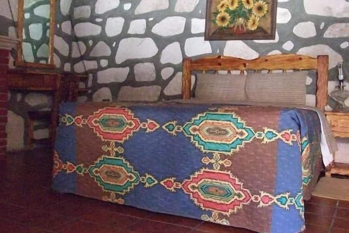 A room with a stone wall, a bed with a patterned bedspread, a wooden headboard, a mirror, and a painting of sunflowers.
