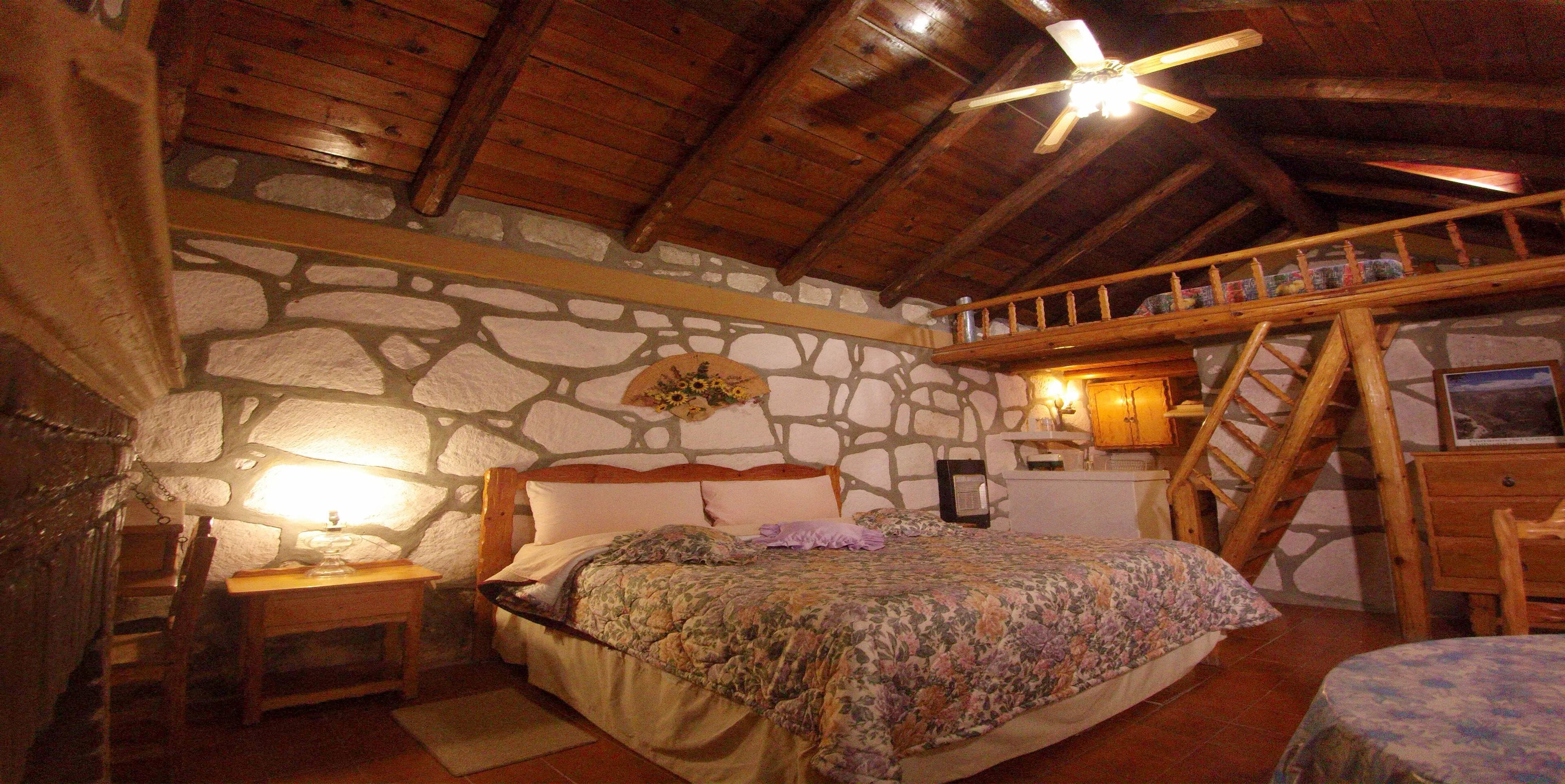 A rustic bedroom with a stone wall, wooden ceiling beams, a bed, a nightstand, a lamp, and a wooden staircase.