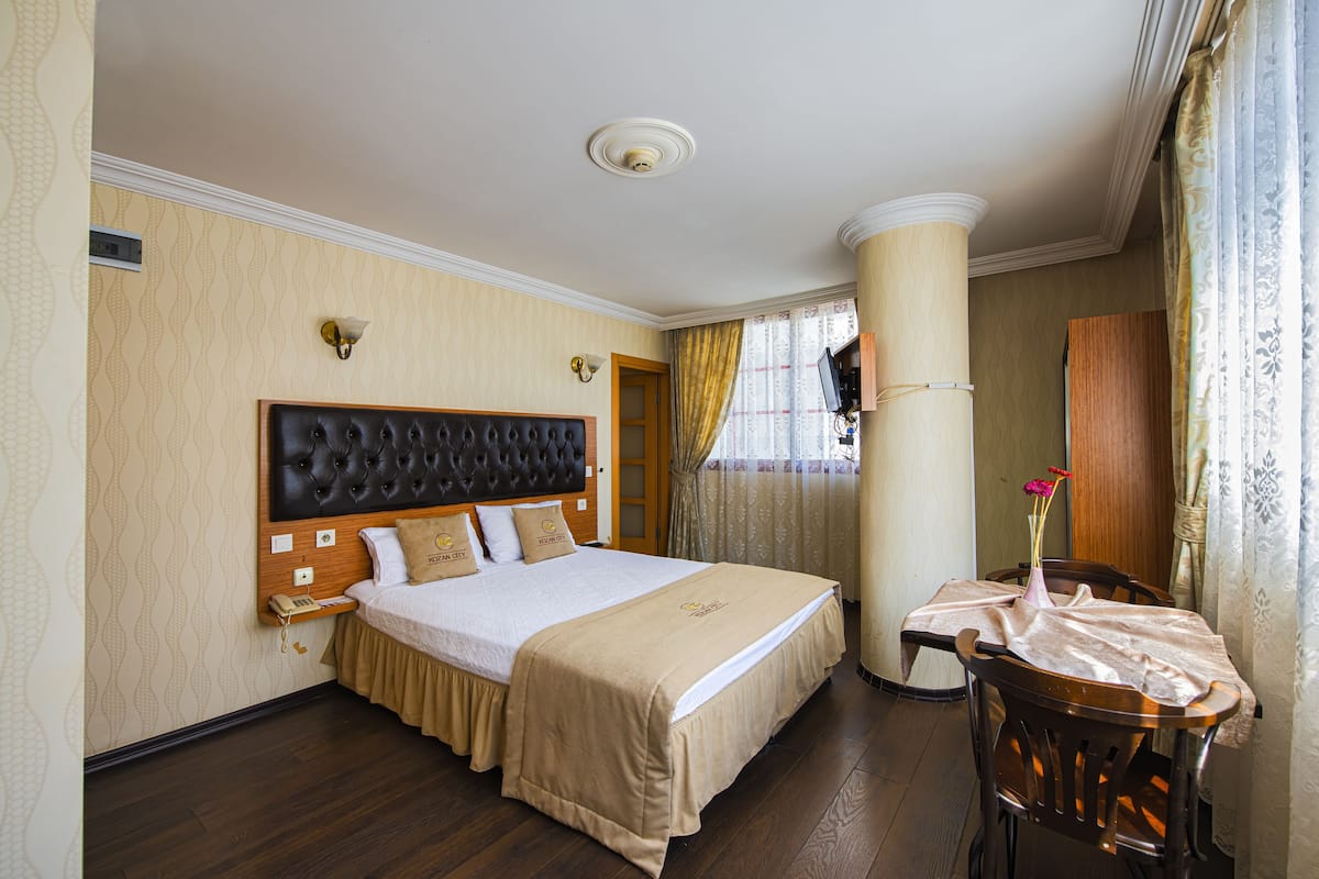Una camera d'albergo con un letto grande, una testiera in legno, un comodino con una lampada, una sedia e una finestra con tende.