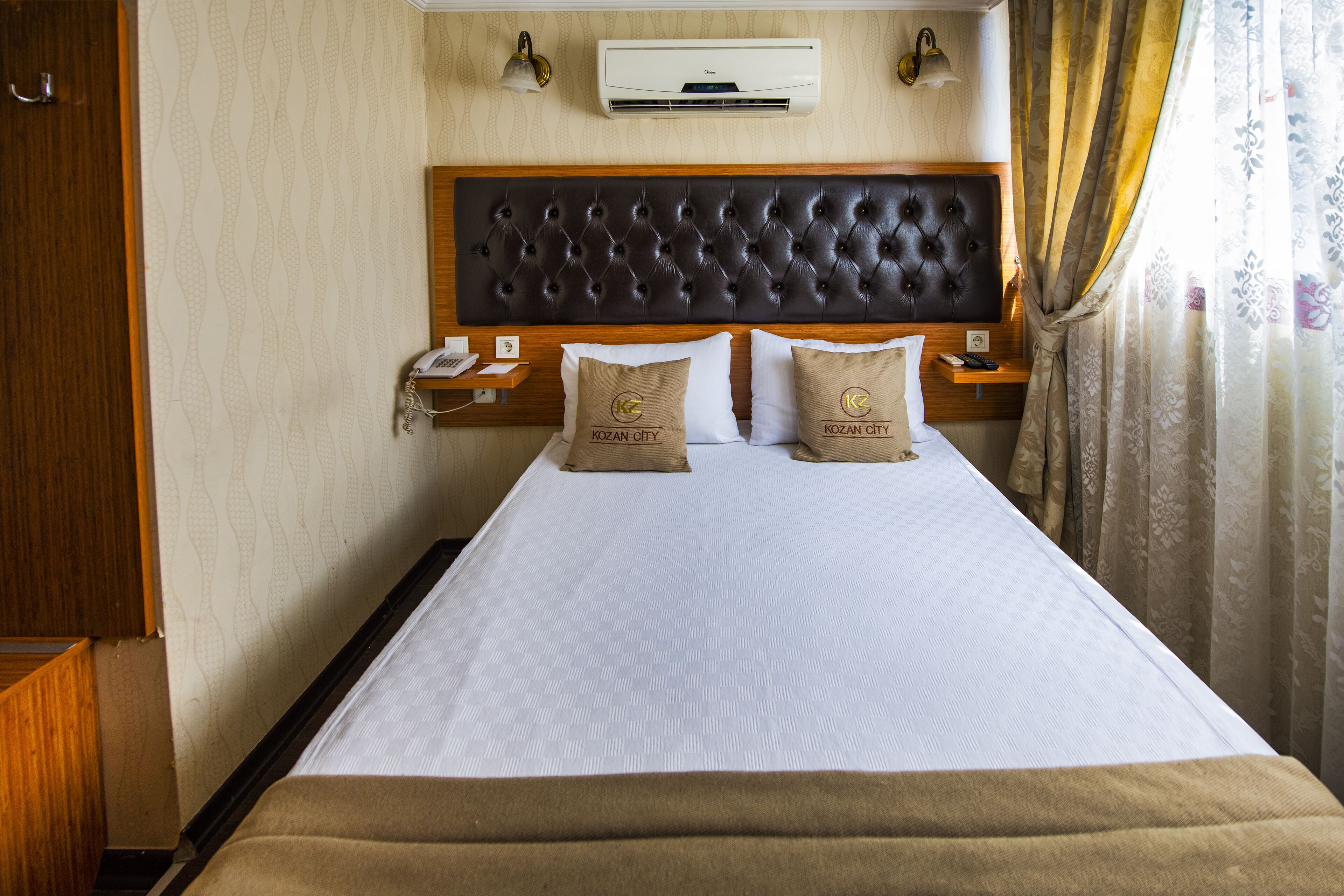 A hotel room with a large bed, a wooden headboard, two pillows with a logo, a bedside table with a lamp, and an air conditioning unit.