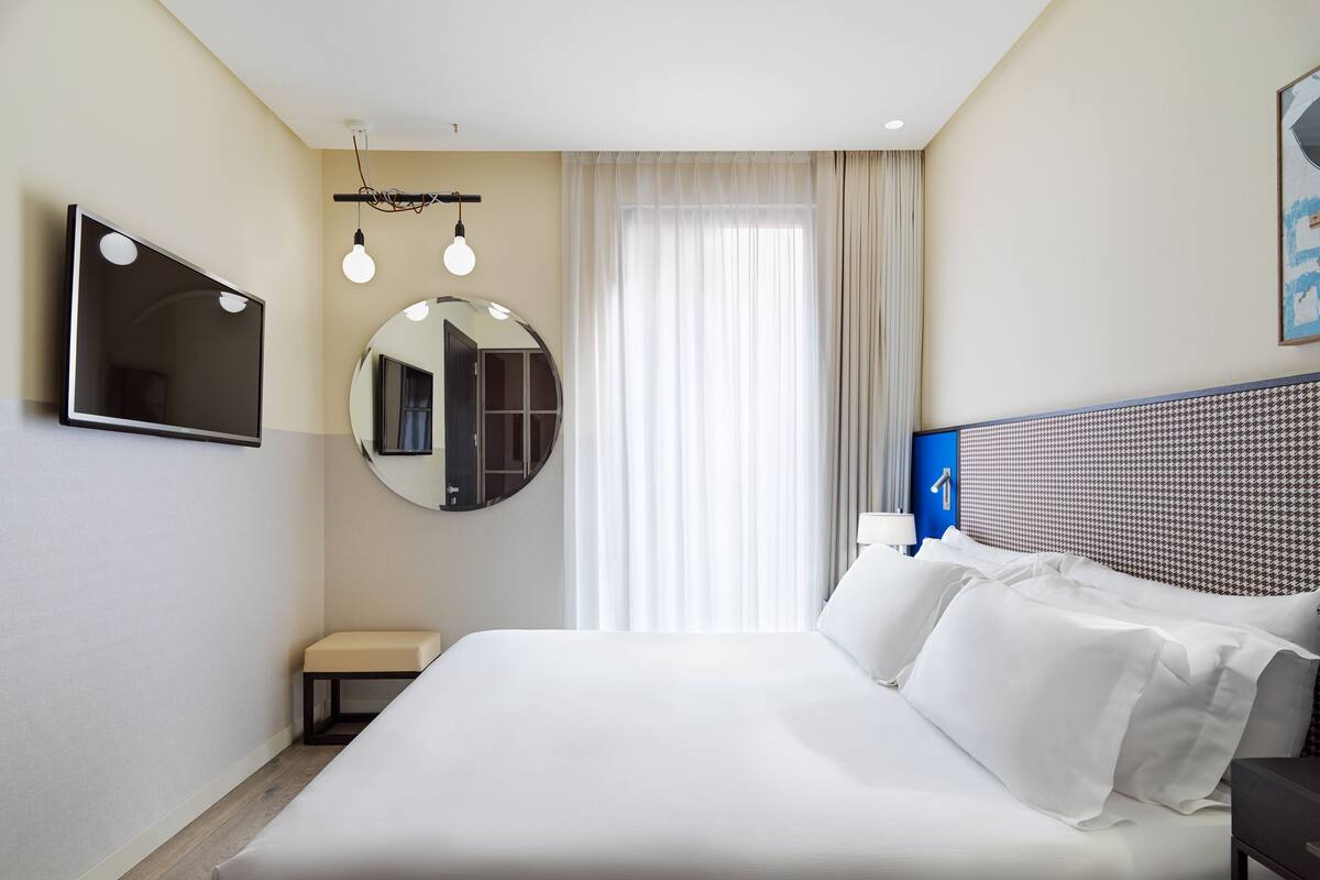 A hotel room with a large bed, a round mirror, a flat-screen TV, and a window with curtains.
