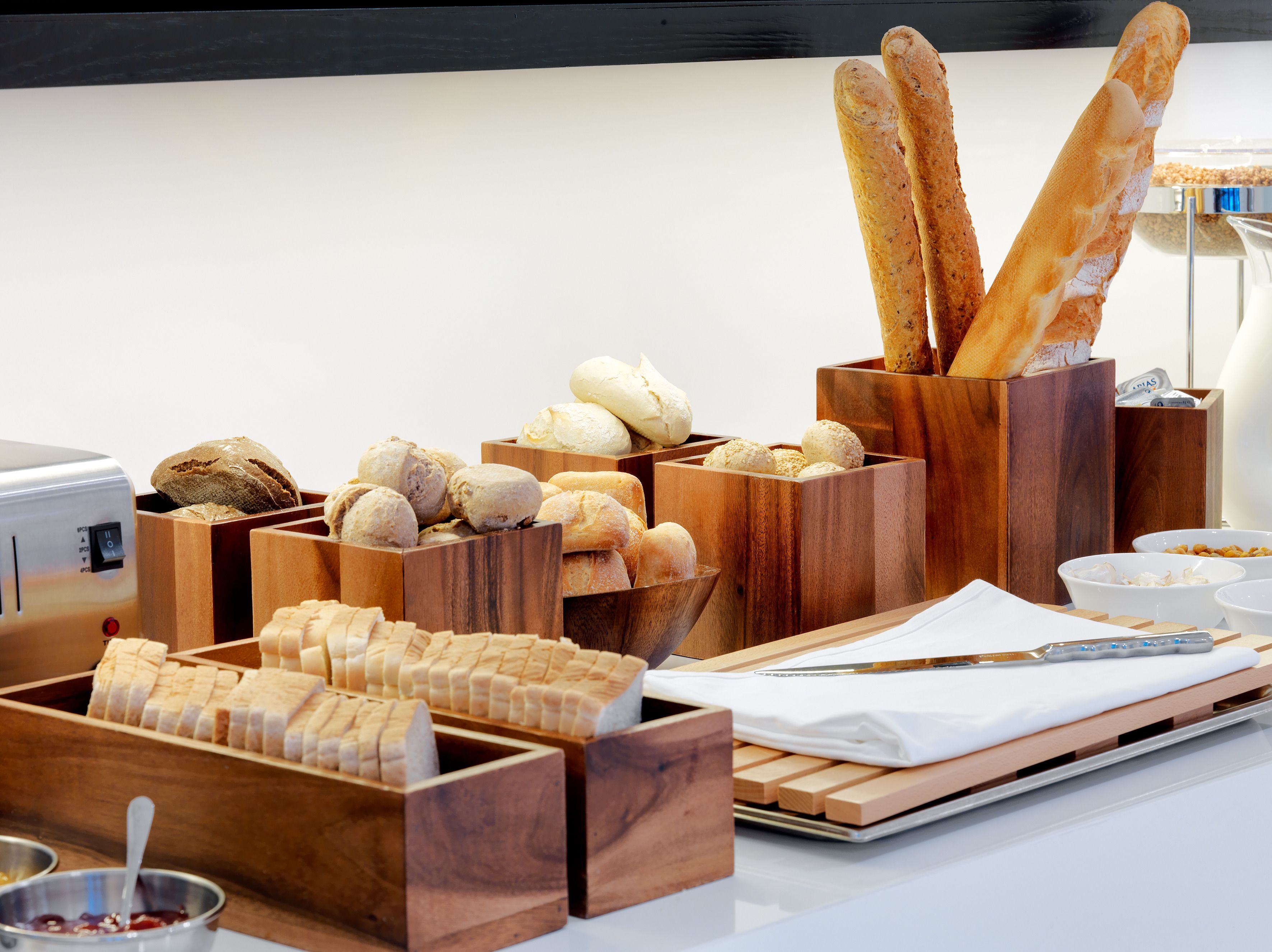 A variety of breads and pastries in wooden containers, a knife, and a bowl of jam on a table.