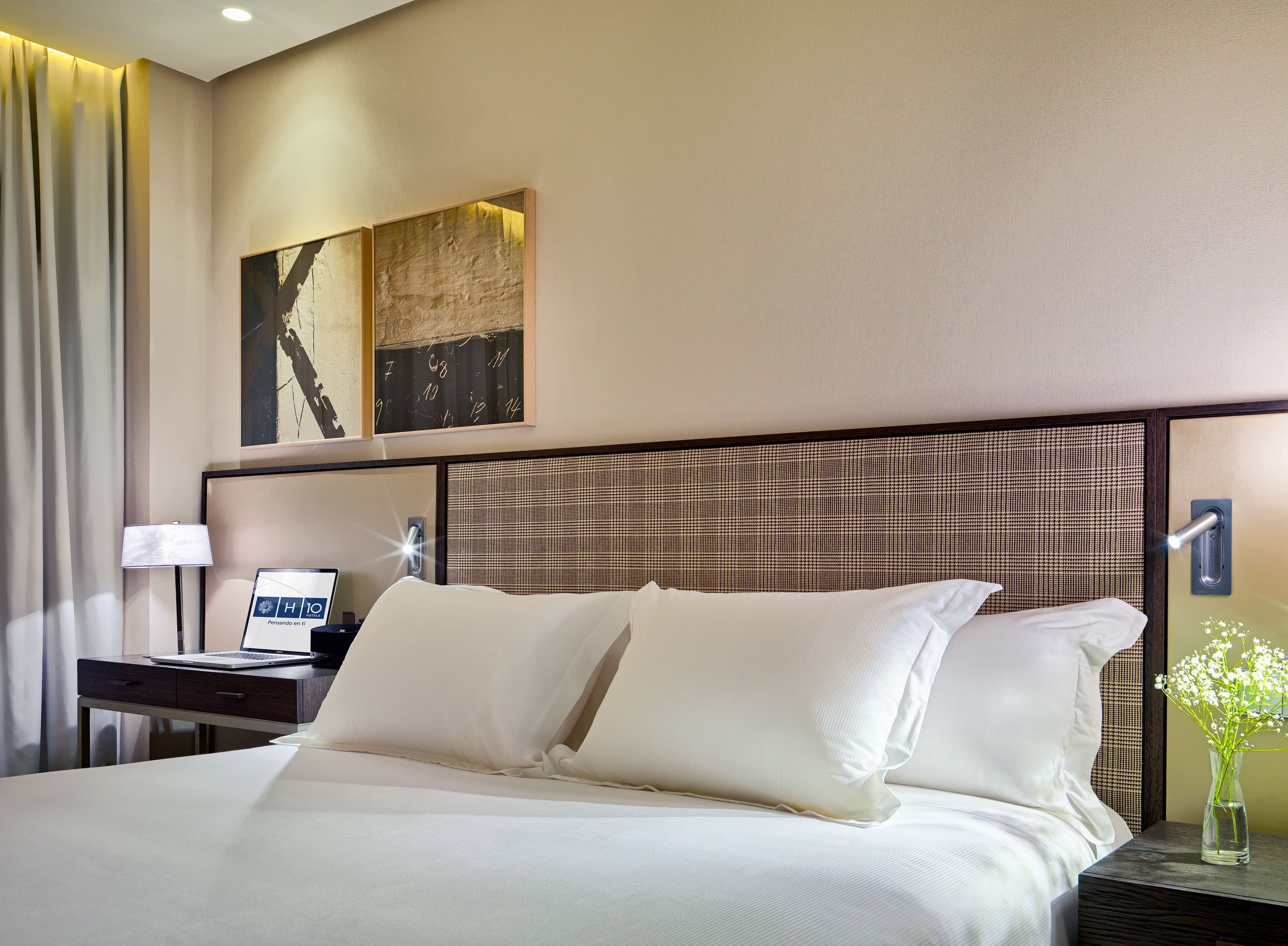 Una habitación de hotel con una cama grande, un escritorio con una computadora portátil, una mesita de noche con un florero y un reloj de pared.