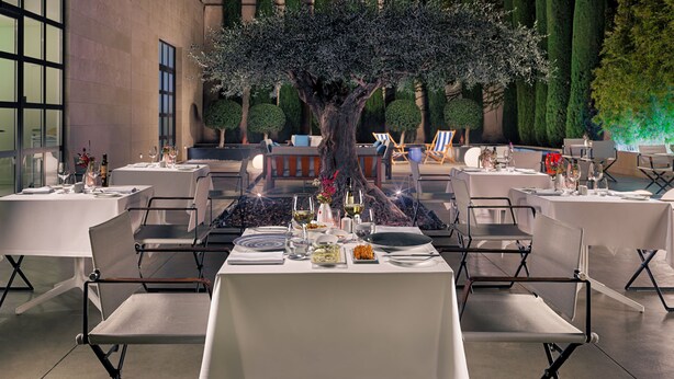 Een restaurant met gedekte tafels, een grote boom in het midden en een uitzicht op de gevel.