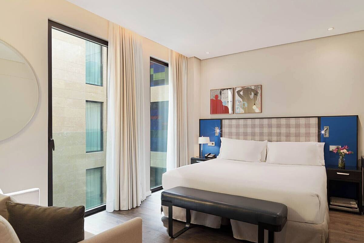 A hotel room with a large bed, a bench, a desk with a lamp, and a view through a glass door.