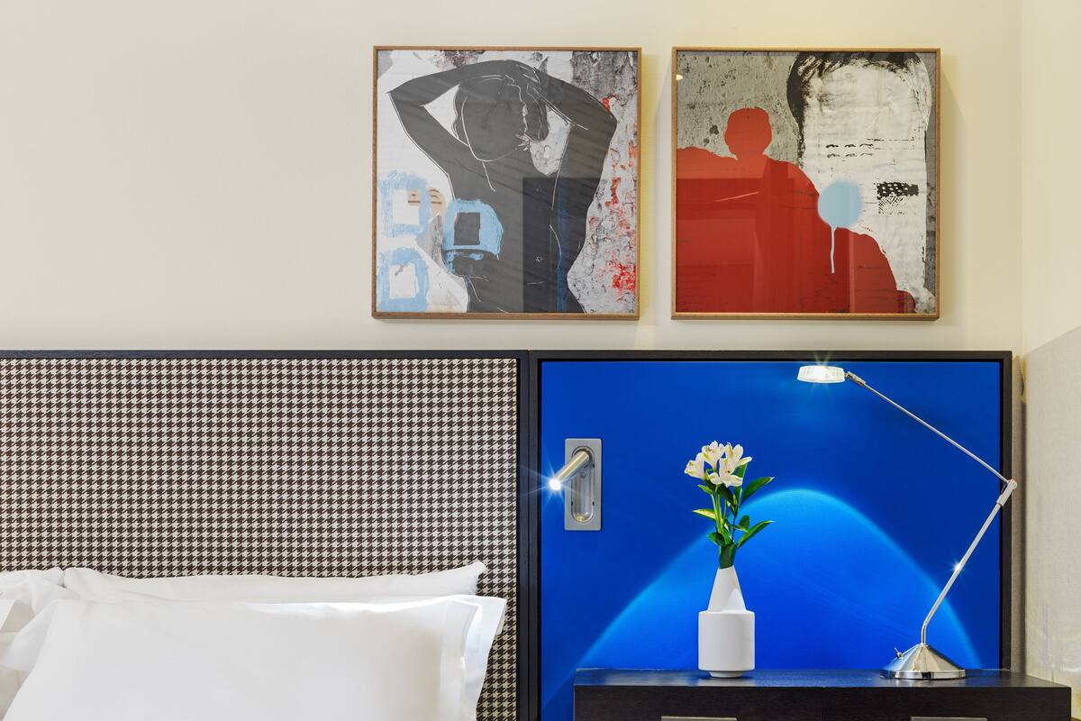 A hotel room with a bed, a desk with a lamp, a vase with flowers, and two artworks on the wall.