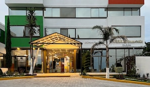 Entrada de un hotel con una zona de espera techada, un cartel que dice 'KISANO SUI Suites' y el reflejo de palmeras en el fachada de vidrio.