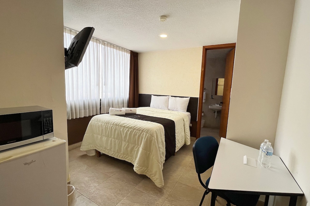 Habitación de hotel con cama, escritorio, silla, microondas y baño visible a través de una puerta abierta.