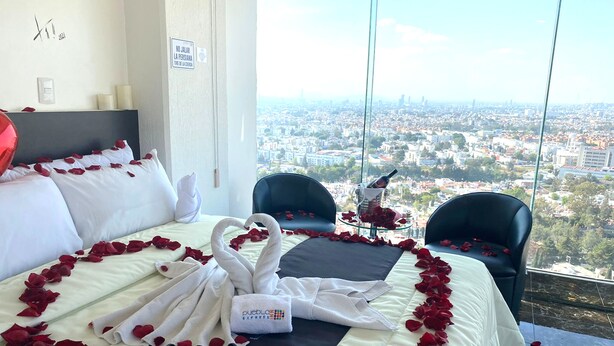 Habitación de hotel con una cama decorada con pétalos de rosa y cisnes, vista a la ciudad y una mesa de vidrio con una botella y rosas.