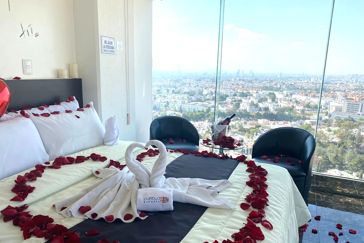 Habitación de hotel con una cama decorada con pétalos de rosa y cisnes, vista a la ciudad y una mesa de vidrio con una botella y rosas.