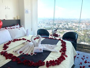 A hotel room with a bed adorned with rose petals and swans, a city view, and a glass table with a bottle and roses.