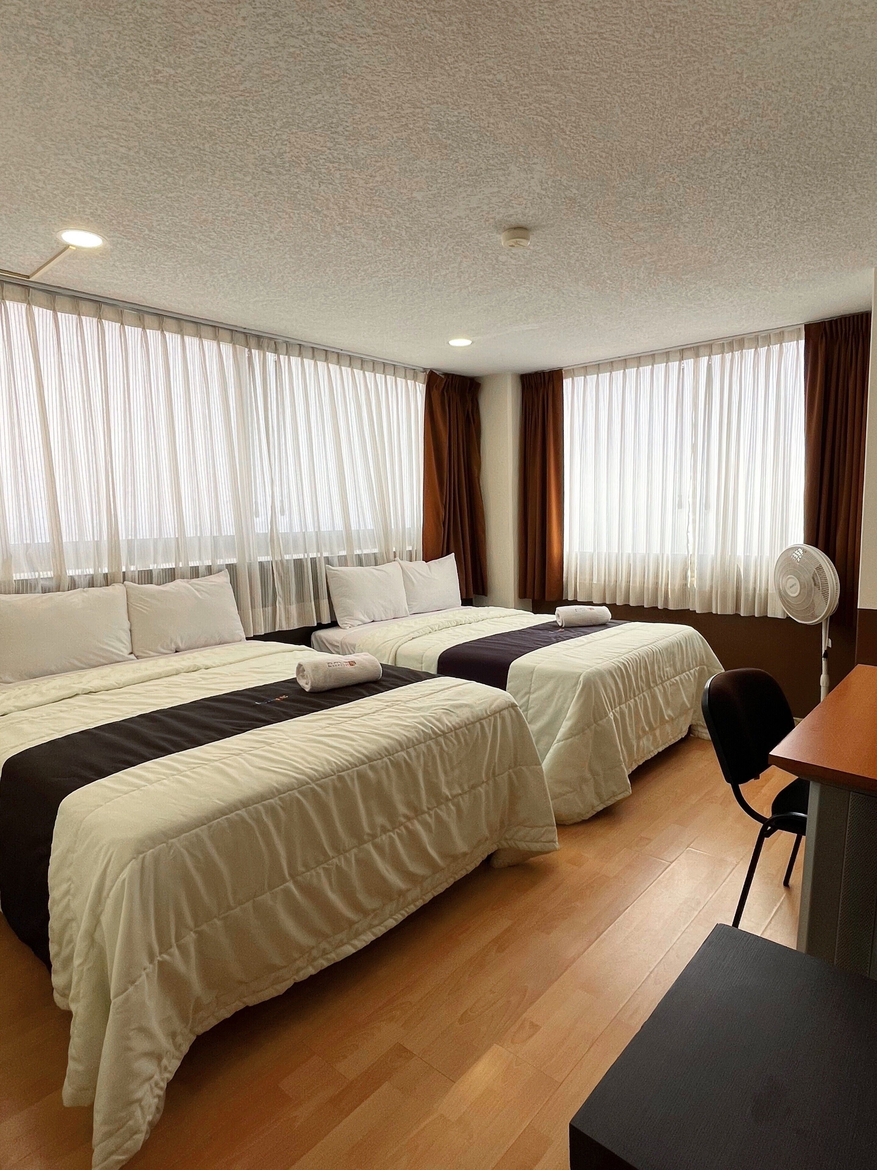 A hotel room with two beds, a desk, a chair, and a fan.