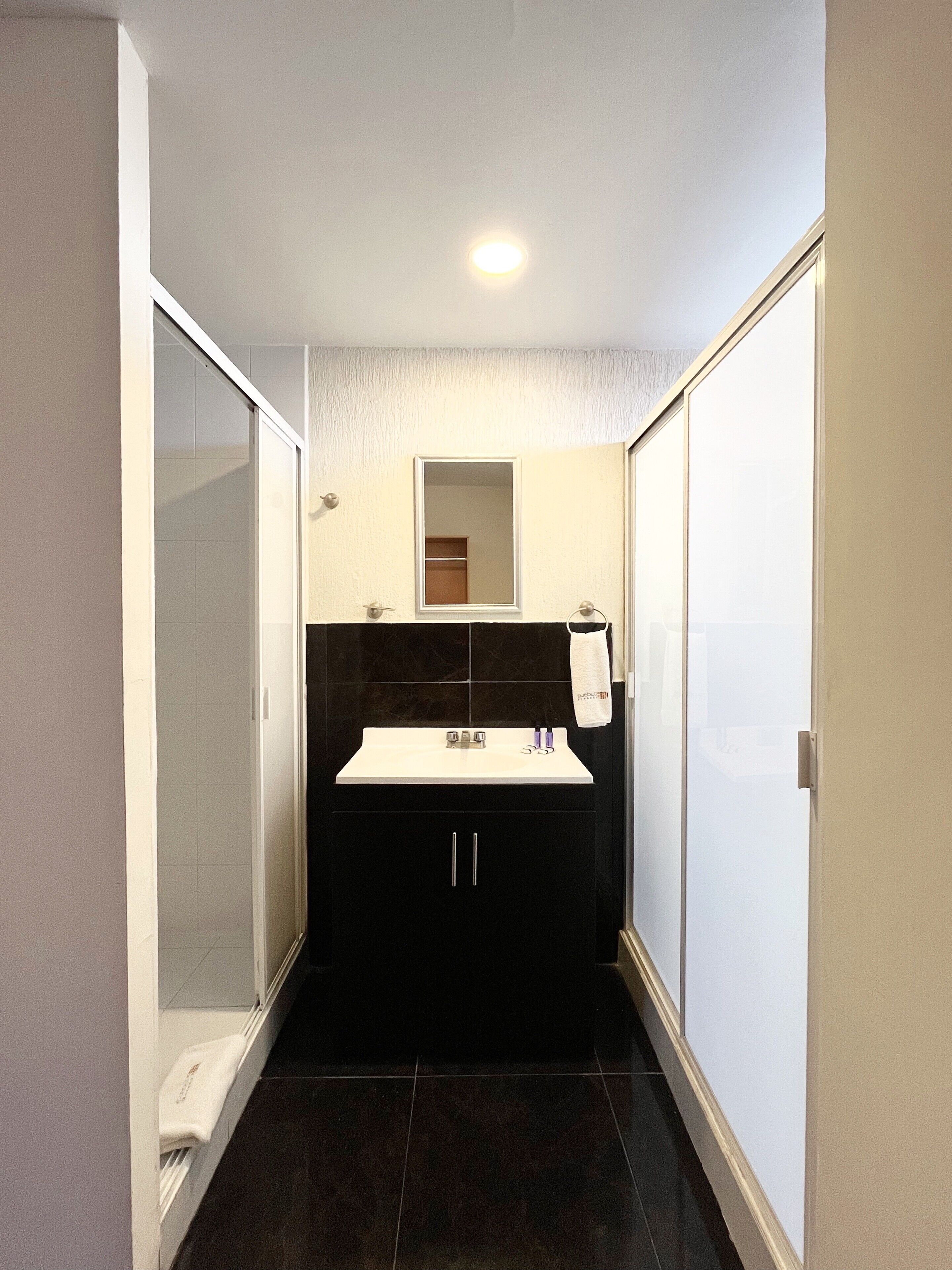 Een kleine badkamer met een wit wastafelmeubel, donkere tegels, een glazen douchecabine en een spiegel boven het wastafelmeubel.