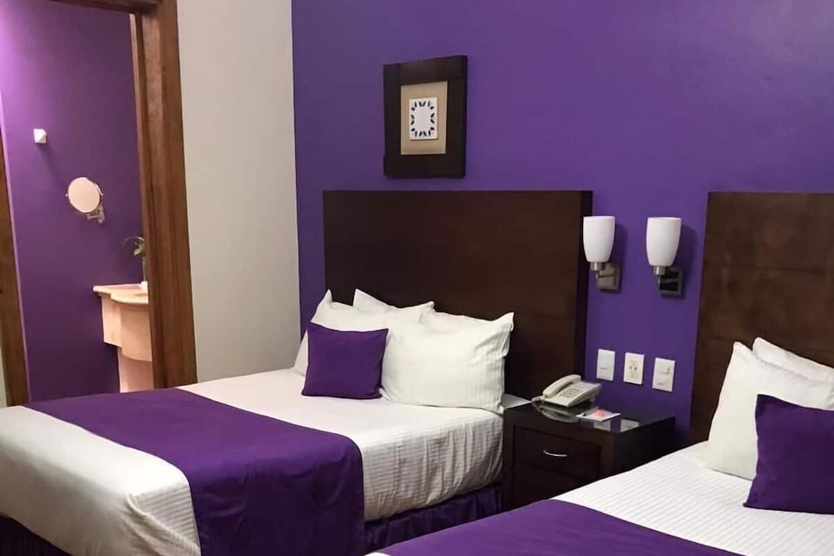 A hotel room with two beds, each with a purple bedspread and white pillows, a wooden headboard, a nightstand with a phone, and a wall-mounted clock.