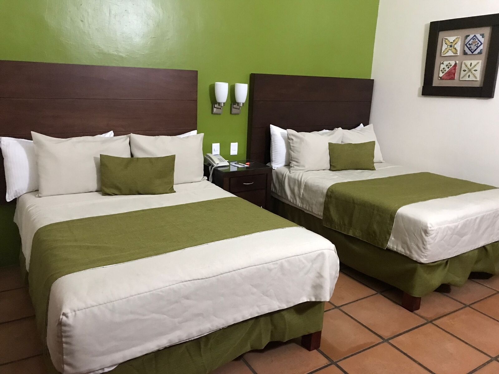 Habitación de hotel con dos camas, cada una con ropa blanca y almohadones verdes, una mesita de noche con teléfono, y una pared verde con un cuadro enmarcado.