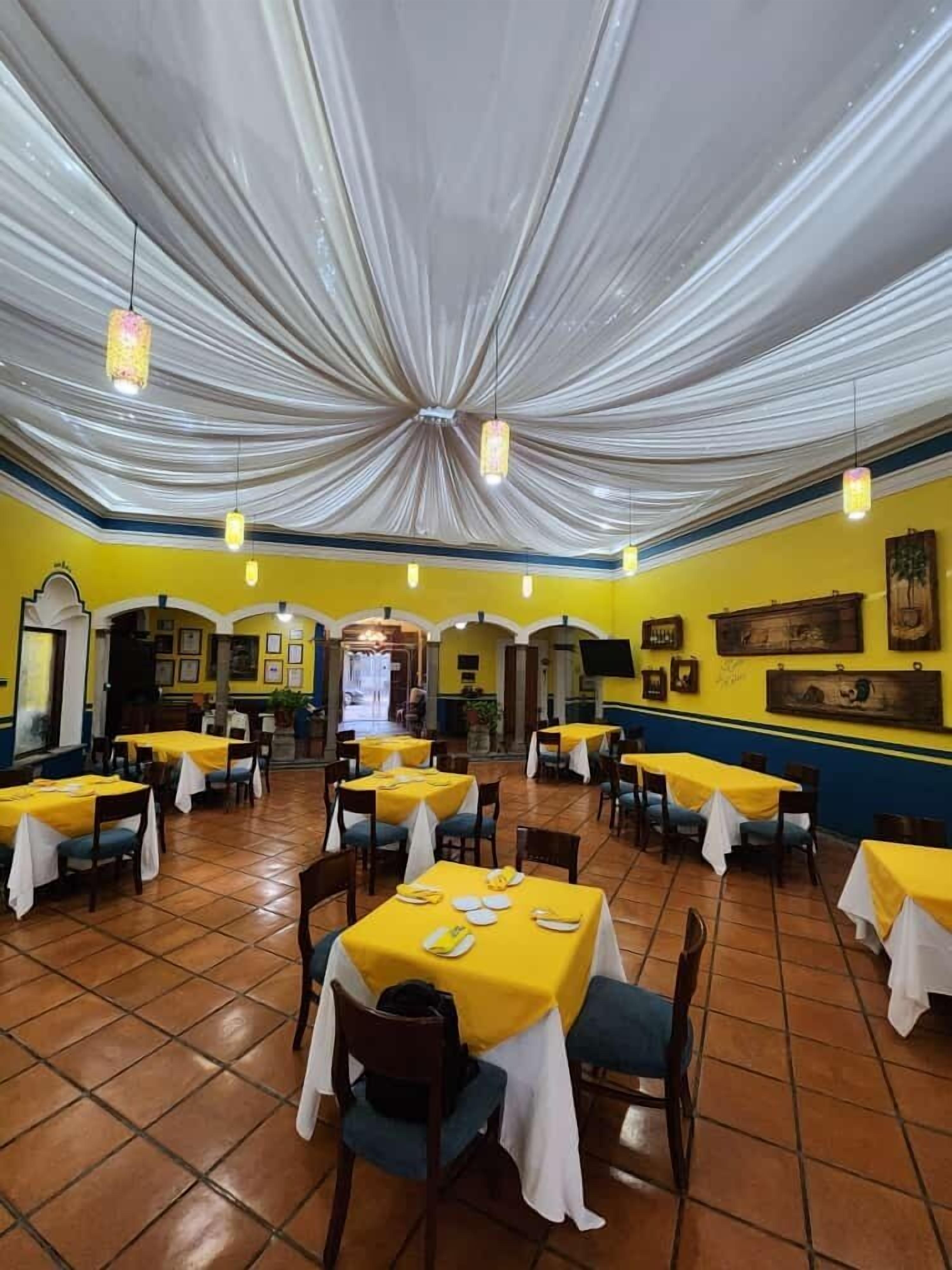 Un ristorante con pareti gialle, tendaggi bianchi a soffitto e numerosi tavoli con tovaglie gialle.