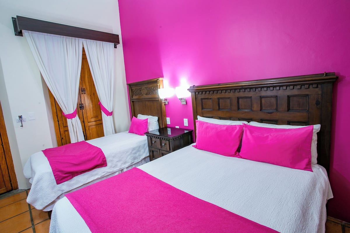 A hotel room with two beds, each with white bedding and pink pillows. There is a wooden headboard on one bed and a nightstand with a mirror and lamp. The room has a bright pink wall and a wooden door with white curtains.