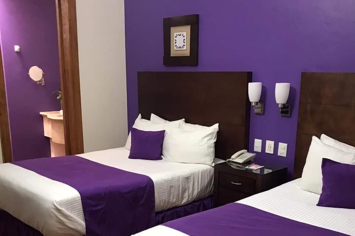 A hotel room with two beds, each with a purple bedspread and white linens. There is a nightstand with a telephone and a small table lamp. The walls are painted purple, and there is a framed picture above the headboard.