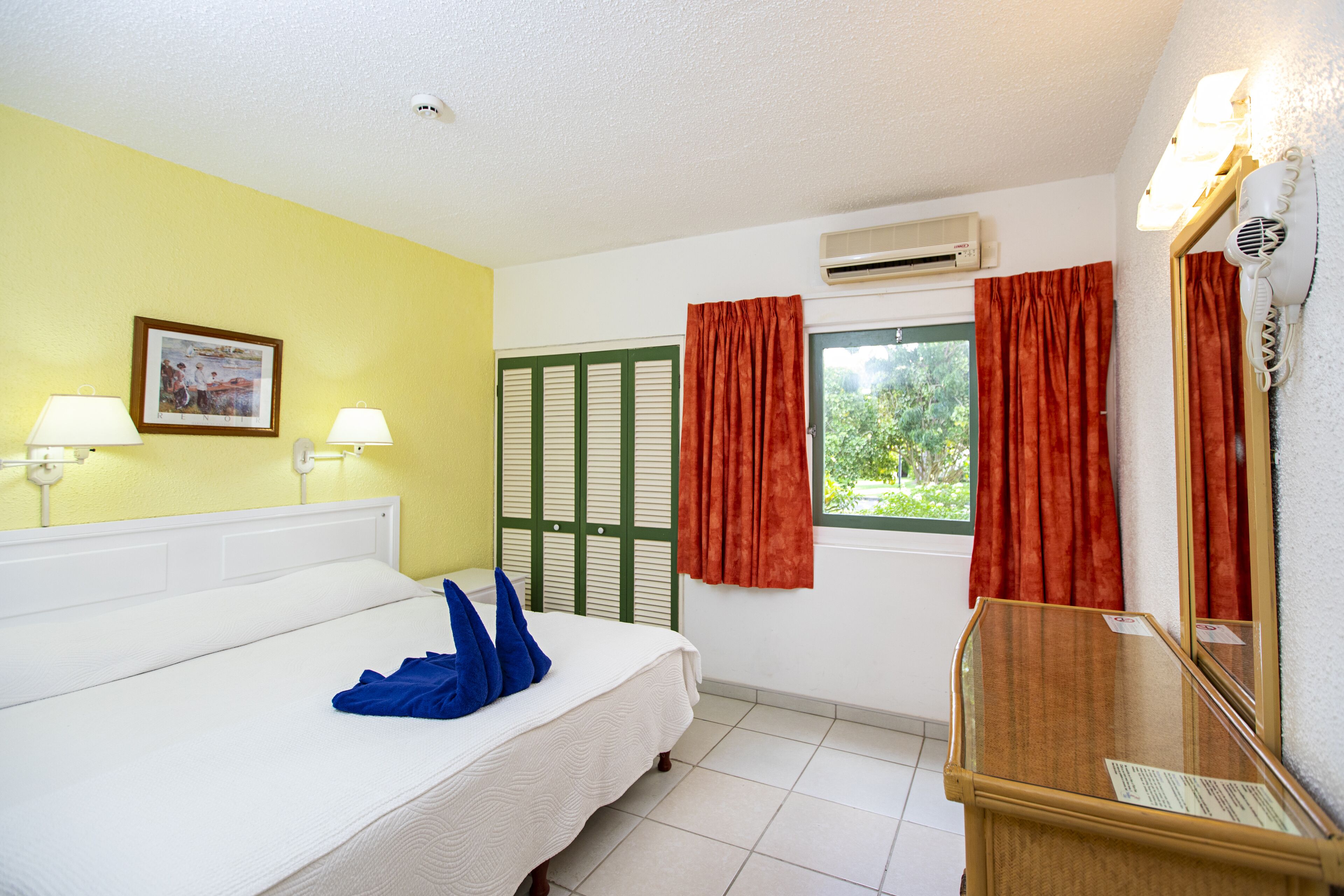 Une chambre d’hôtel comprenant un lit, deux lampes de chevet, une fenêtre avec des rideaux rouges, un appareil de climatisation et un miroir avec un sèche-cheveux.
