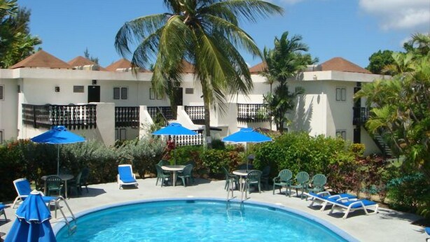Piscina con sillones de descanso y sombrillas, rodeada de palmeras y un edificio con balcones.