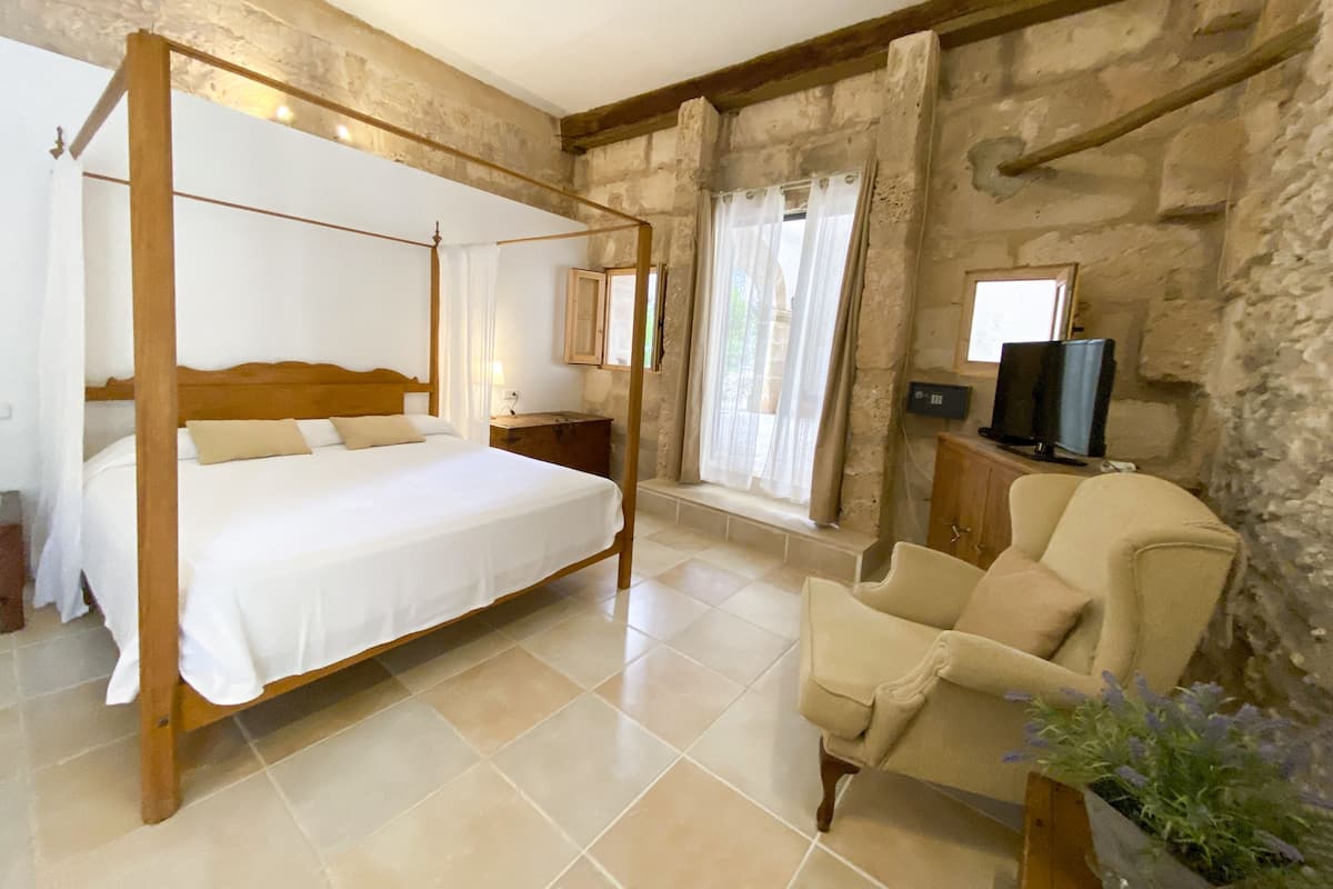Un lit à baldaquin avec une tête de lit en bois, une table de chevet avec une lampe, une chaise et un téléviseur posé sur une commode en bois.
