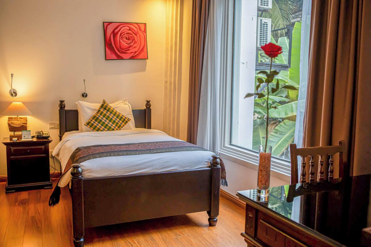 Una cama bien hecha con cabecera, una mesita de noche con lámpara, un cuadro con una rosa y una ventana con vistas a la vegetación.