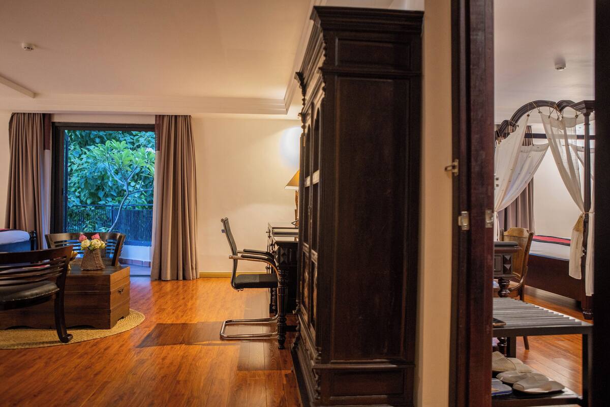 Una habitación con suelo de madera, un mueble alto de madera oscura, una silla, una mesita con un florero y vistas al exterior con vegetación.