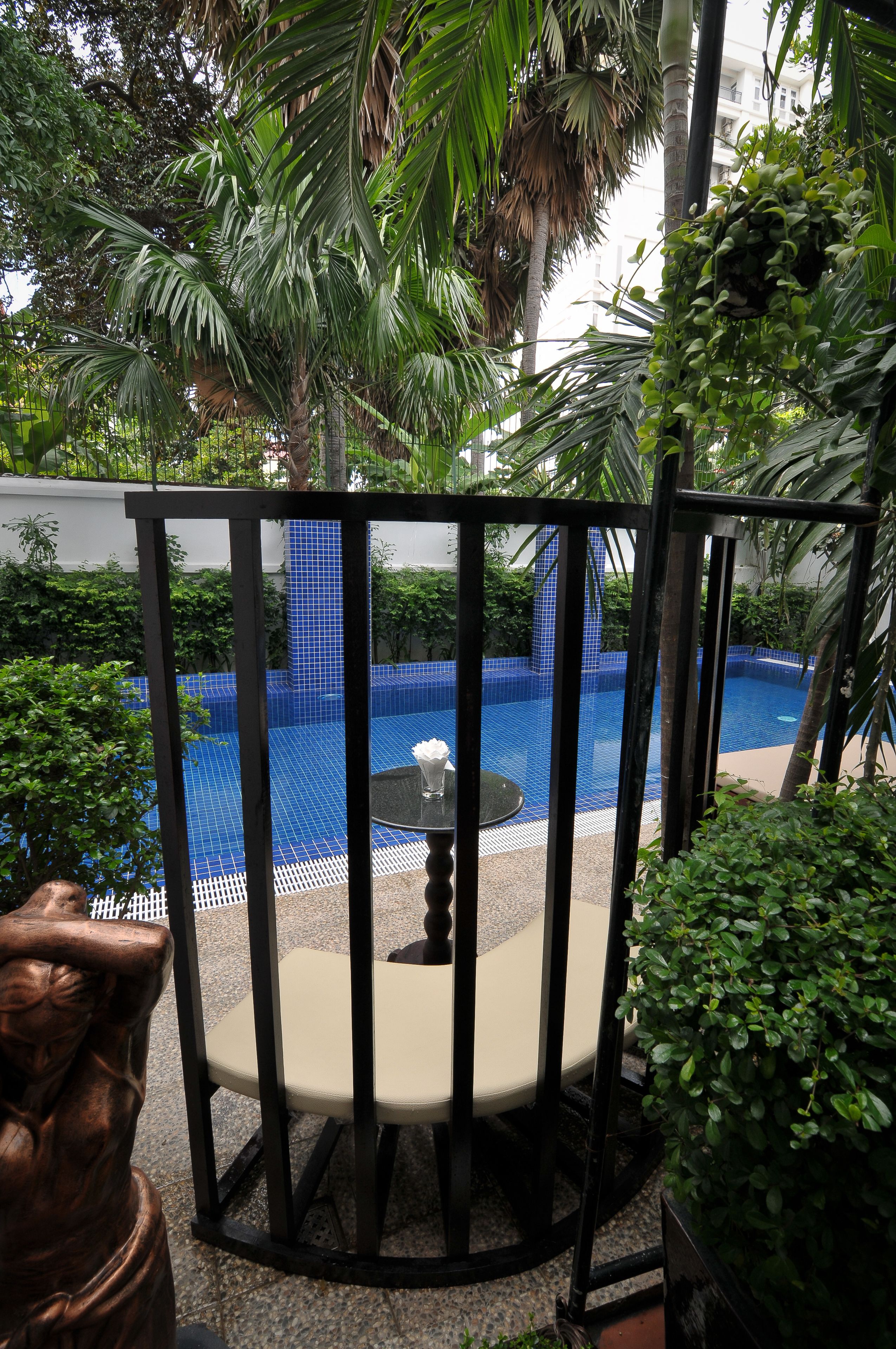 A pool area with a tiled blue pool, surrounded by greenery and a covered seating area with a table and chairs.