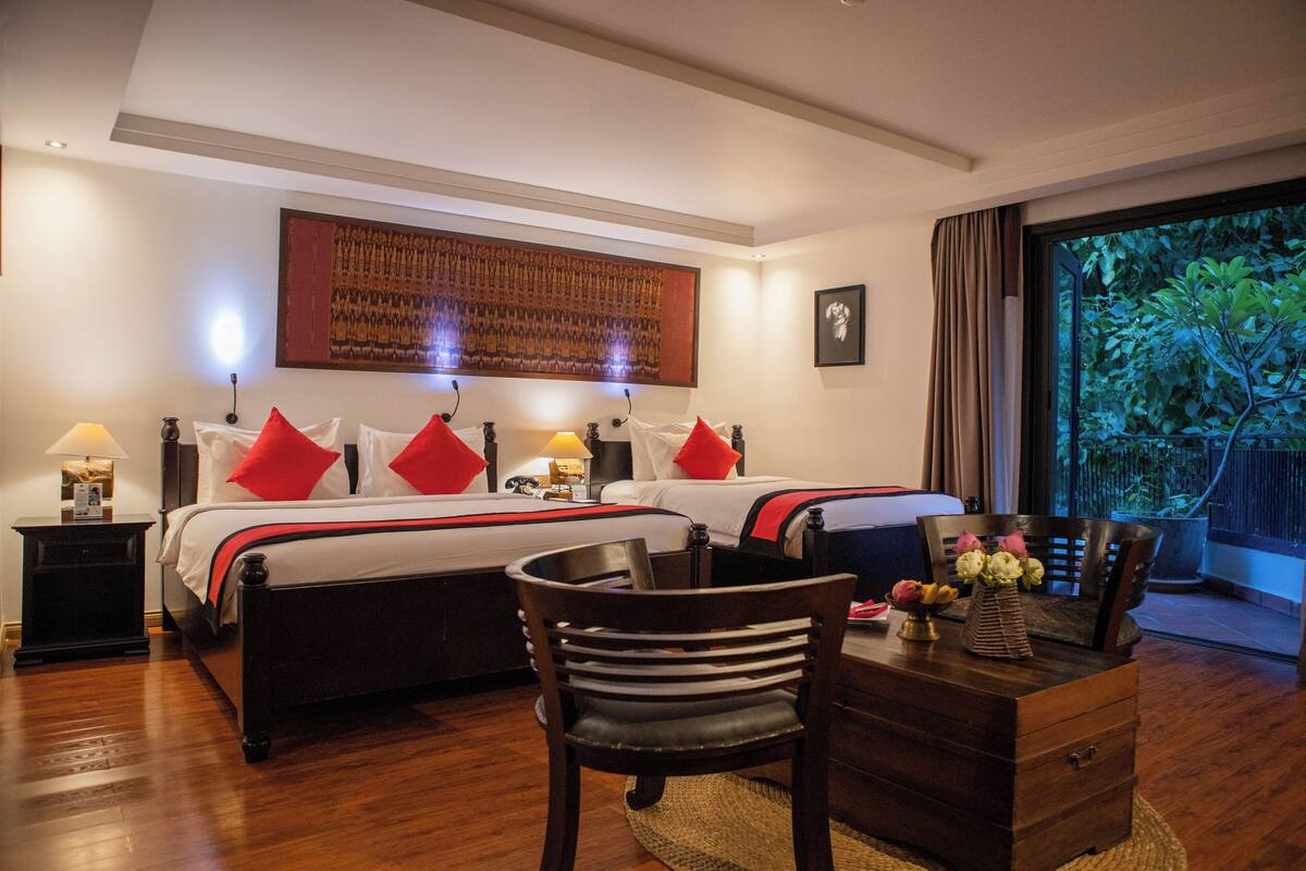 Habitación de hotel con dos camas, un escritorio con silla, una mesita de noche y un ventanal con vistas a la vegetación.