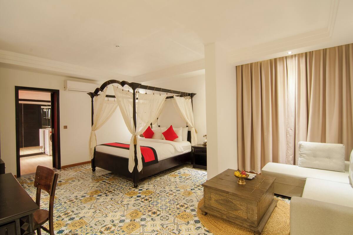 Una cama con dosel con almohadas rojas, una mesa de centro de madera con fruta, un sofá blanco y un suelo estampado.
