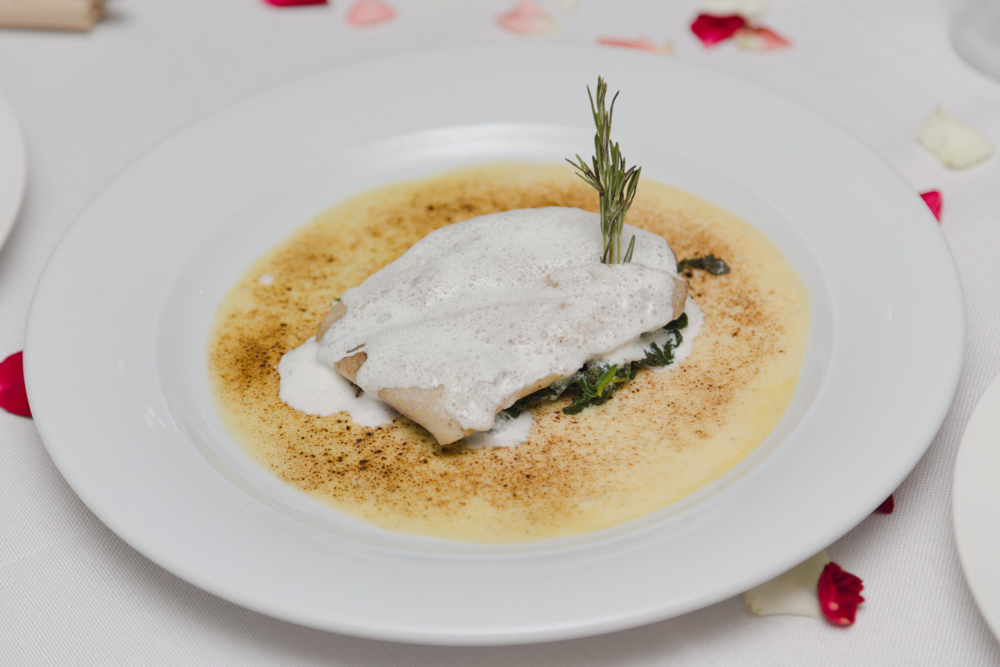 Une assiette blanche sur laquelle est disposé un filet de poisson blanc, agrémenté d’une branche de romarin, le tout servi sur un lit de sauce jaune et saupoudré de poivre rouge.