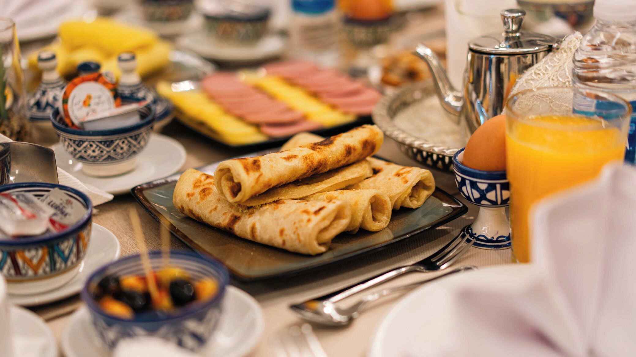 Een ontbijttafel met rolletjes pannenkoeken, een theepot, een schaal fruit en een glas jus d'orange.