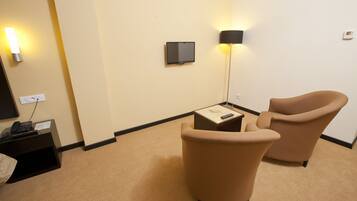 A hotel room with beige carpet, a desk with a telephone, a chair, a TV mounted on the wall, and a floor lamp.