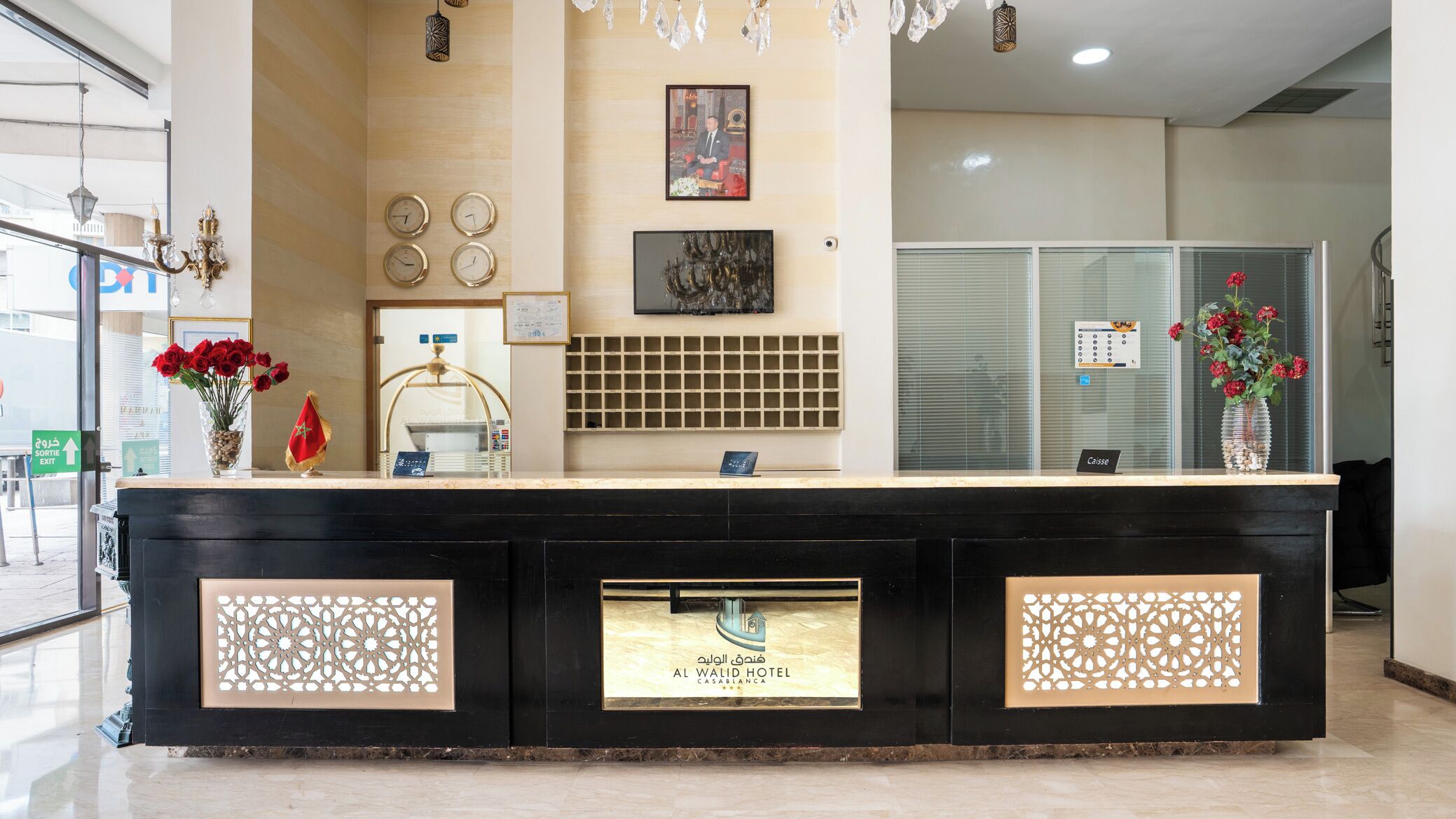 A hotel reception desk with a glass partition, a framed photo above the counter, and a decorative wall clock.