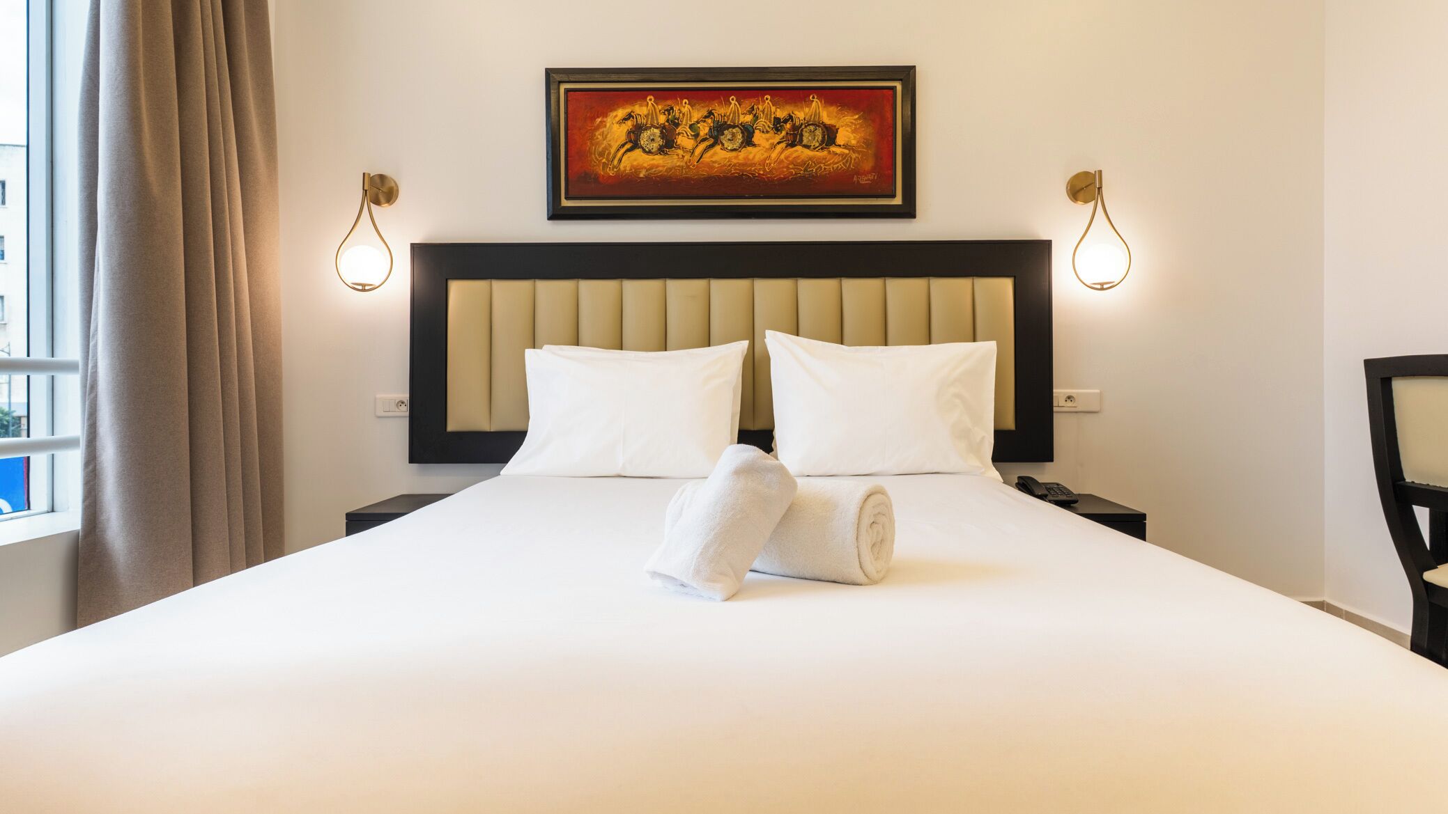A hotel room with a large bed, two bedside tables, a headboard, a painting above the bed, and two wall-mounted lights.