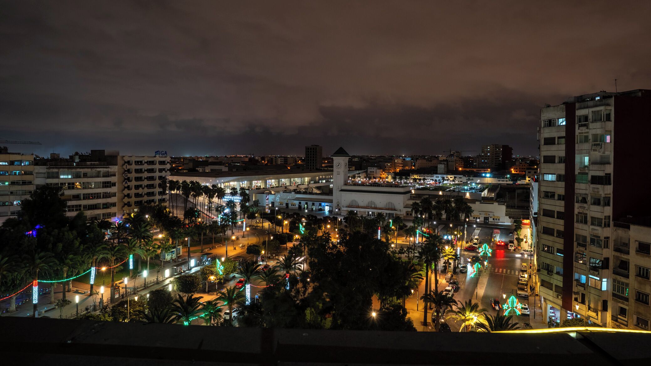 A cityscape at night with illuminated buildings, a prominent clock tower, and a busy street with palm trees.