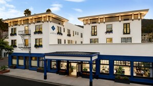Un hotel con fachada blanca, detalles azules y varias ventanas. Cuenta con mesas al aire libre y un área cubierta.