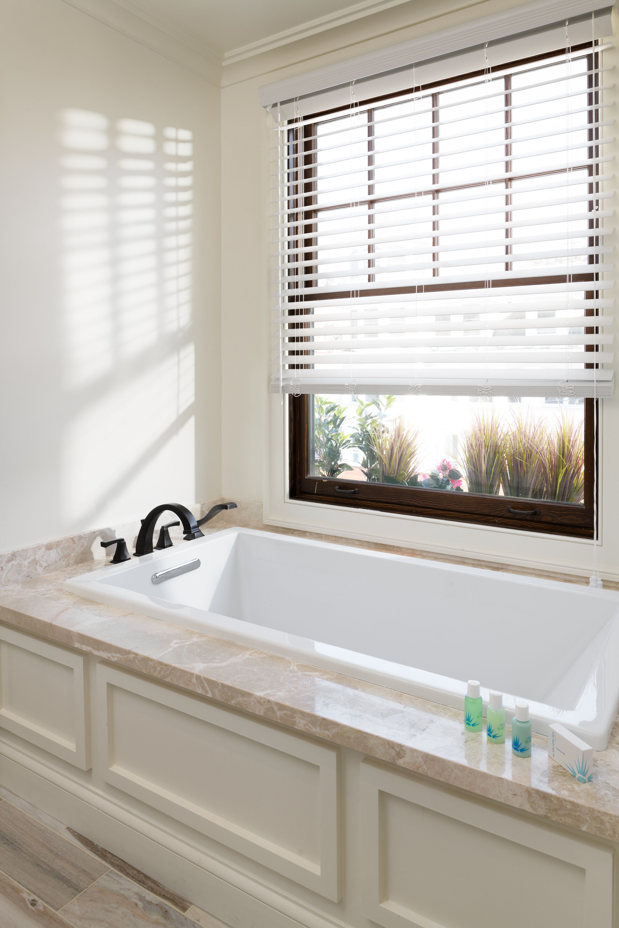 A bathroom with a large white bathtub, a window with blinds, and a marble countertop.