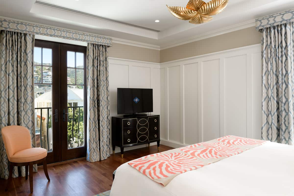 A hotel room with a bed, a television on a dresser, a chair, and a balcony with patterned curtains.