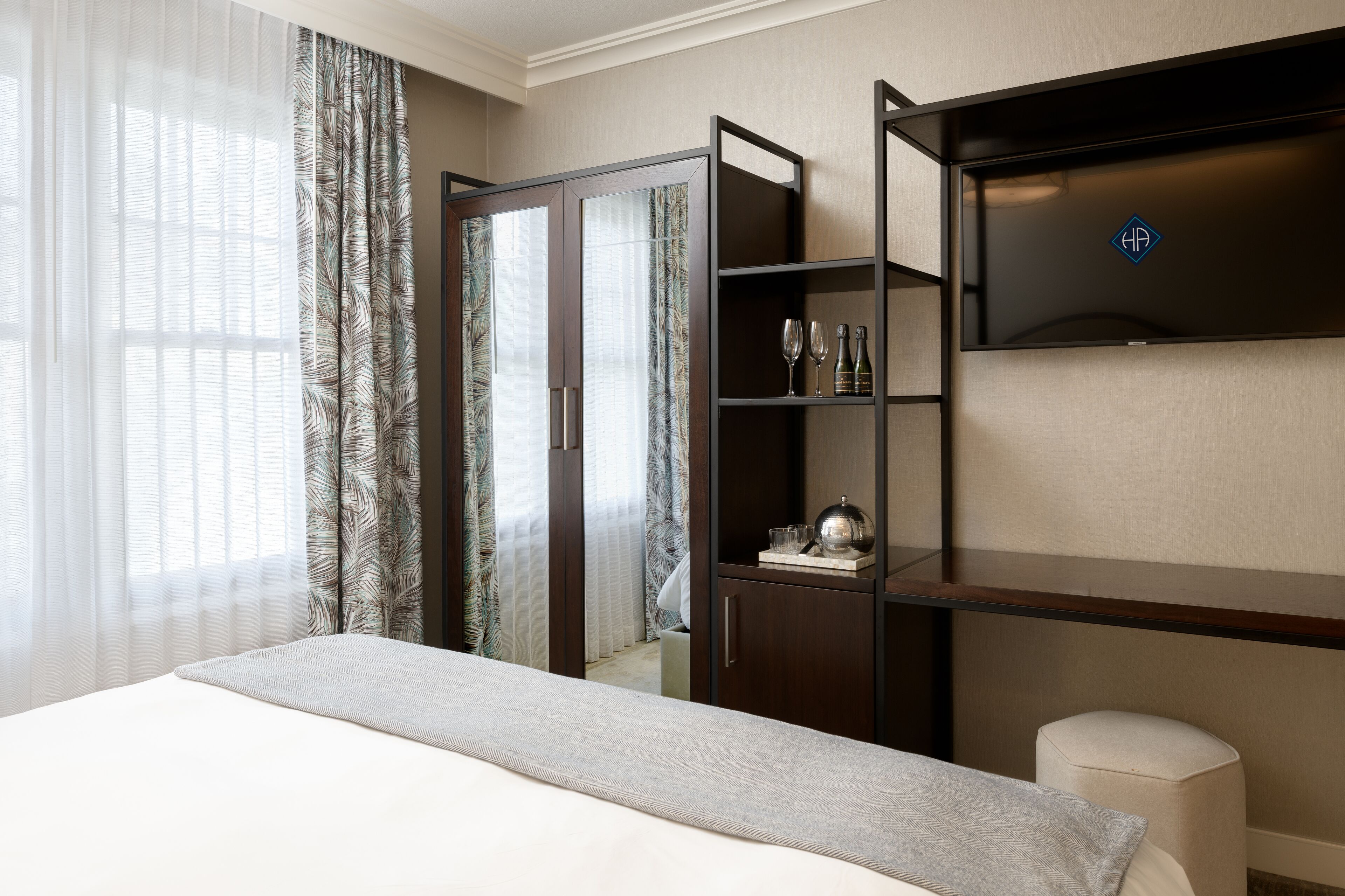 A hotel room with a bed, a wardrobe, a shelf with glasses and a bottle, and a small stool.