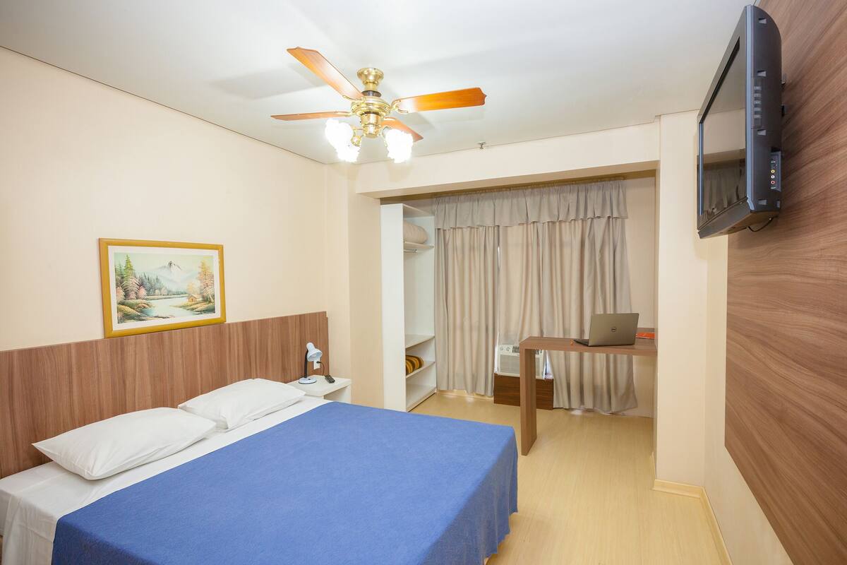 Una habitación de hotel con cama, un escritorio con una computadora portátil, un televisor, un ventilador de techo y un cuadro en la pared.