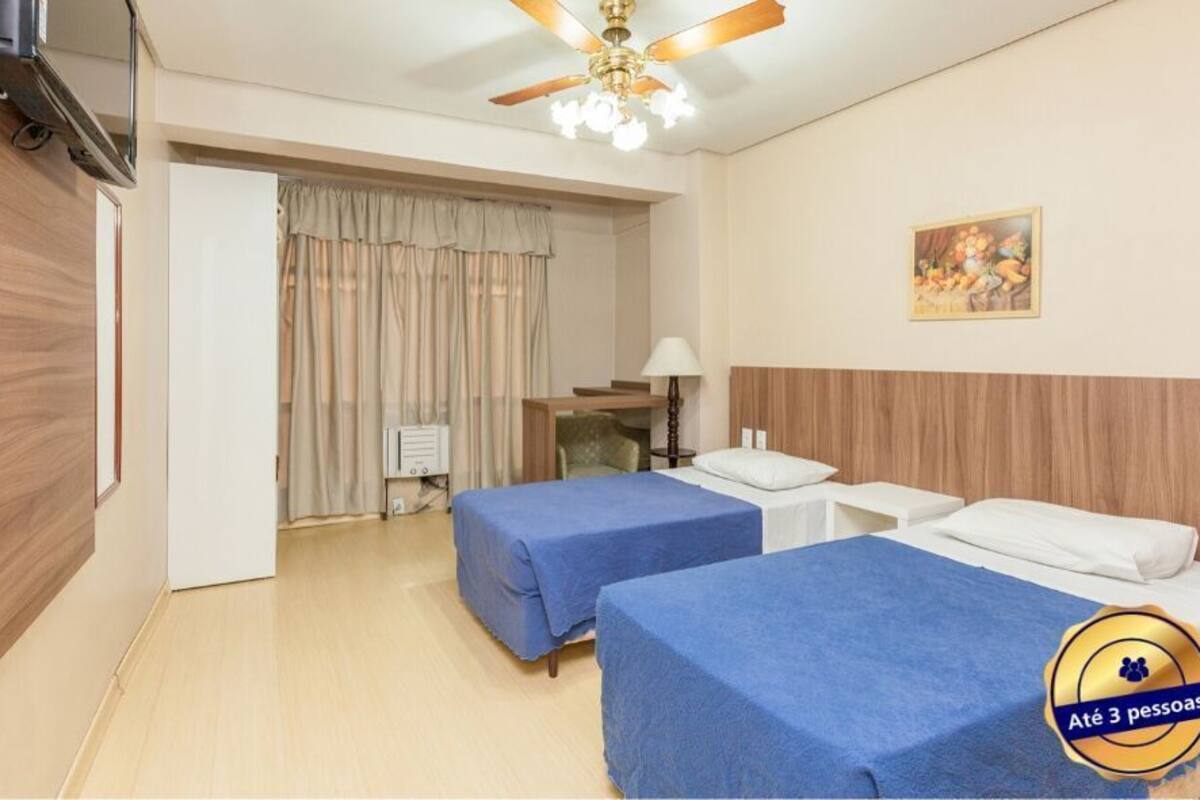 Habitación de hotel con dos camas individuales, ventilador de techo, televisión y un cuadro en la pared.