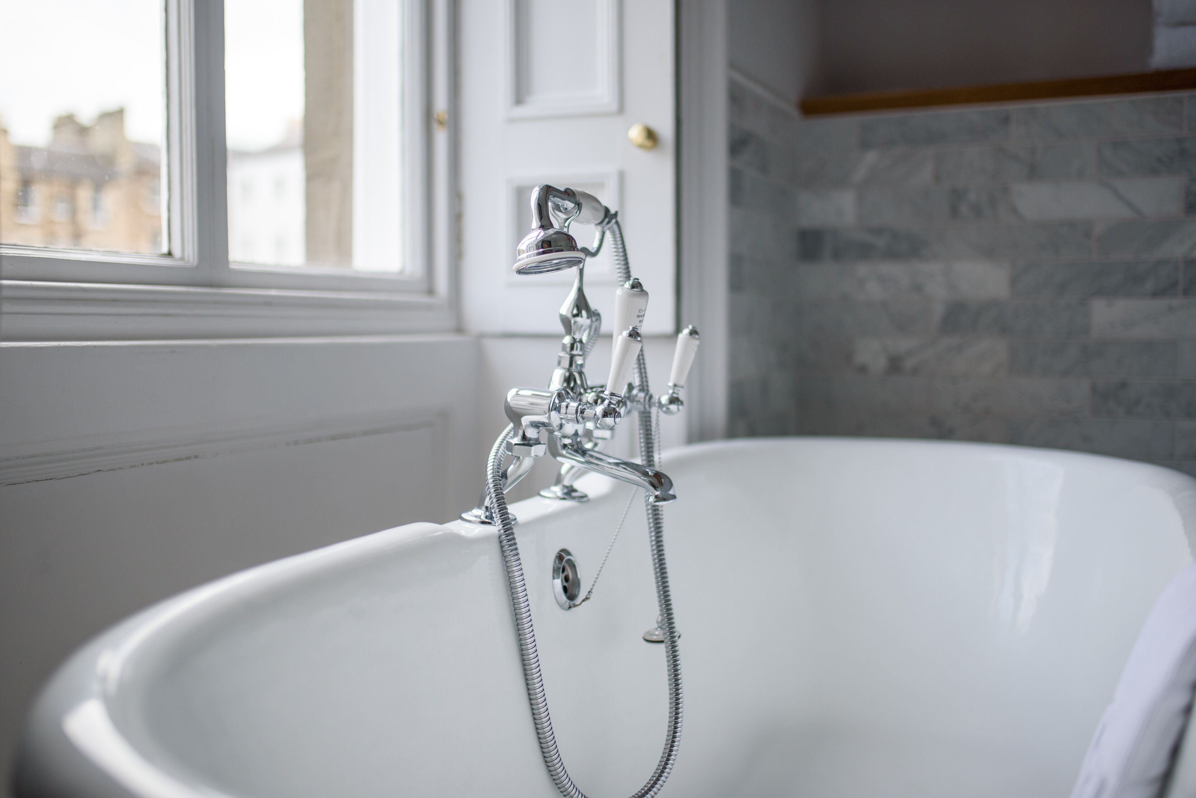 A modern bathroom with a freestanding bathtub, a visible showerhead, and a window providing a view of buildings outside.