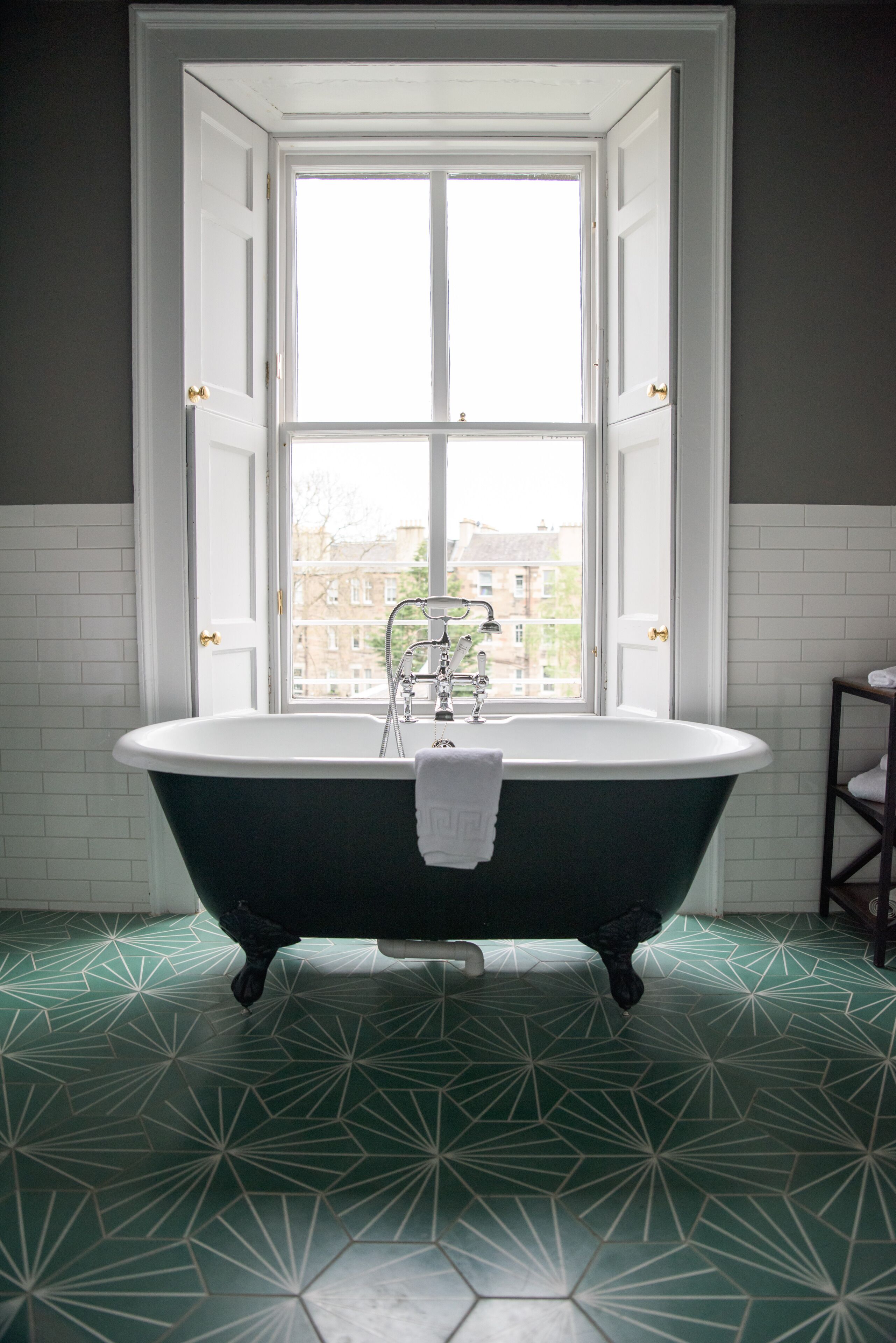 A bathroom with a freestanding bathtub, a window with a view of buildings, and a patterned floor.