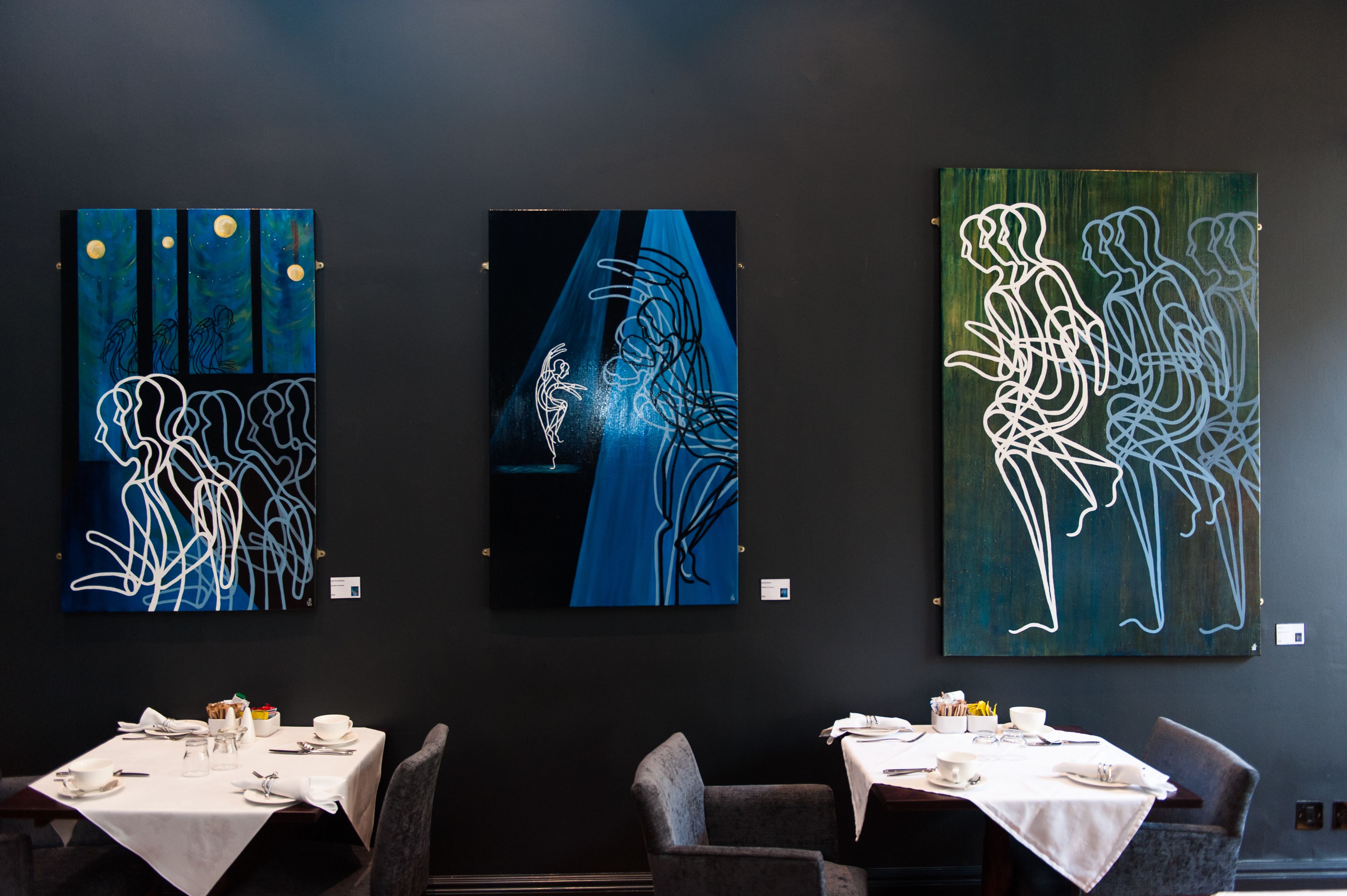 Three artworks on a wall in a dining area with tables set for dining.