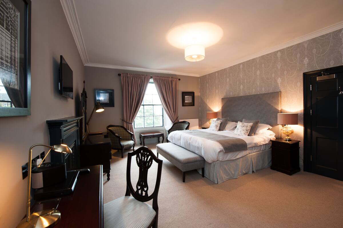 A hotel room with a large bed, a desk with a lamp, a chair, a television, and a window with curtains.