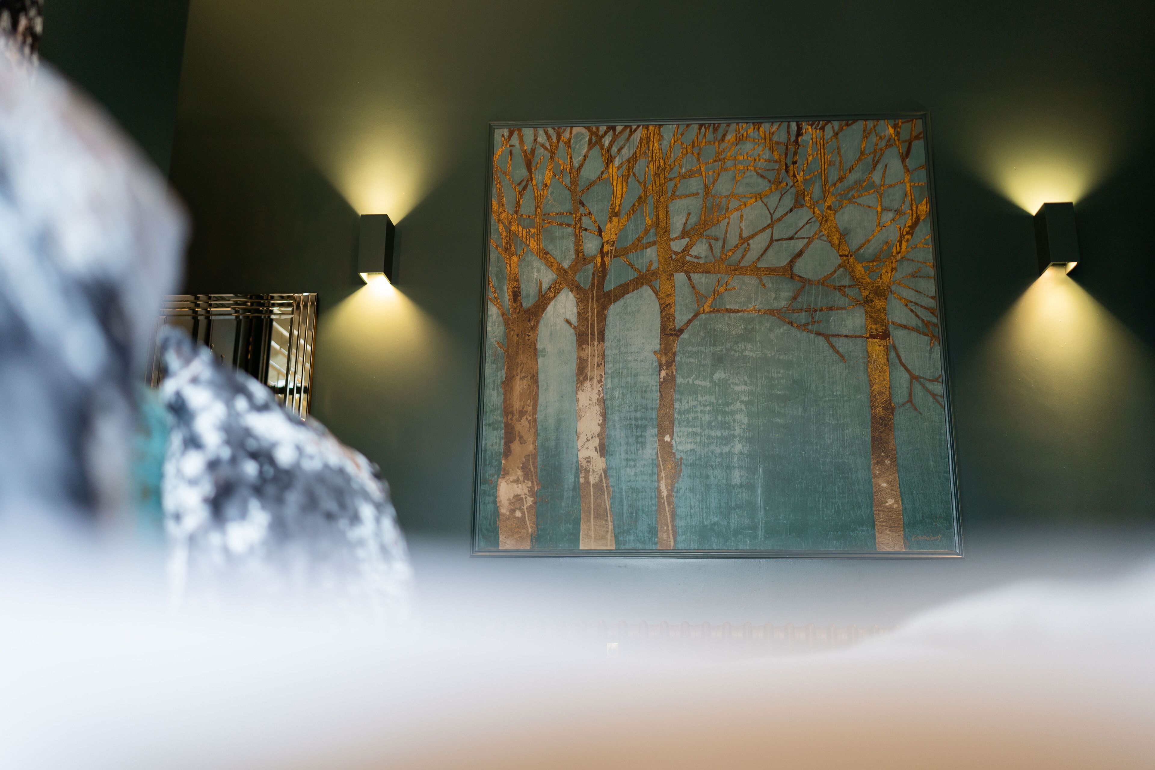 A framed artwork of bare trees on a wall, with two wall-mounted lights on either side.