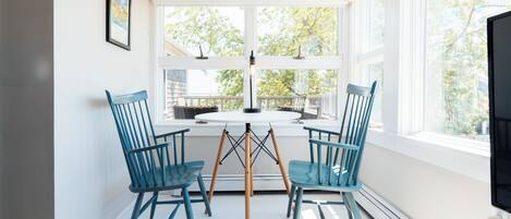 A dining area with a round table and four blue chairs, large windows with a view of trees, and a television on a stand.