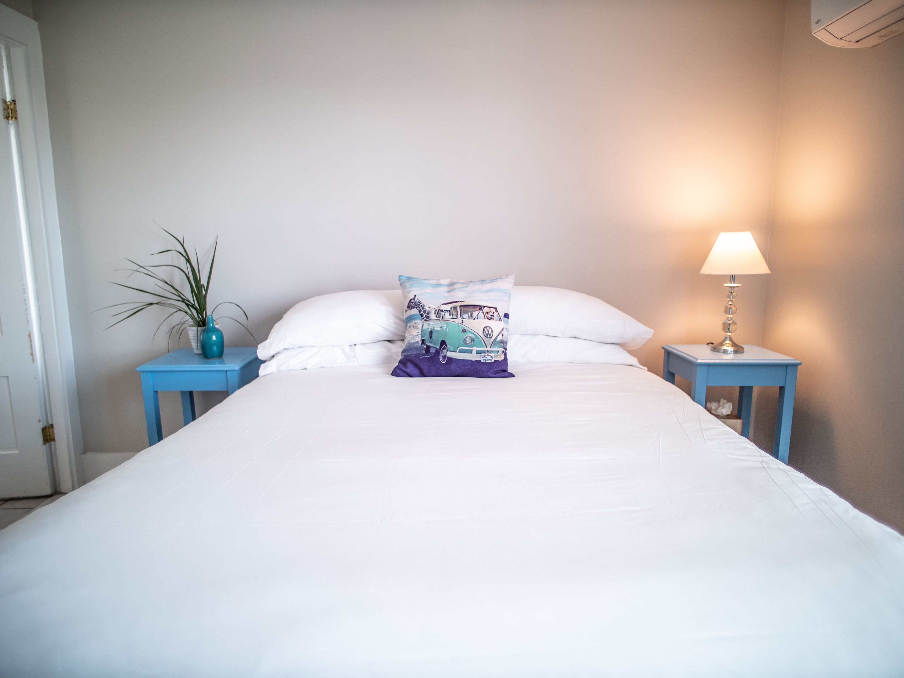 A neatly made bed with white bedding, a blue bedside table with a lamp, and a decorative pillow.