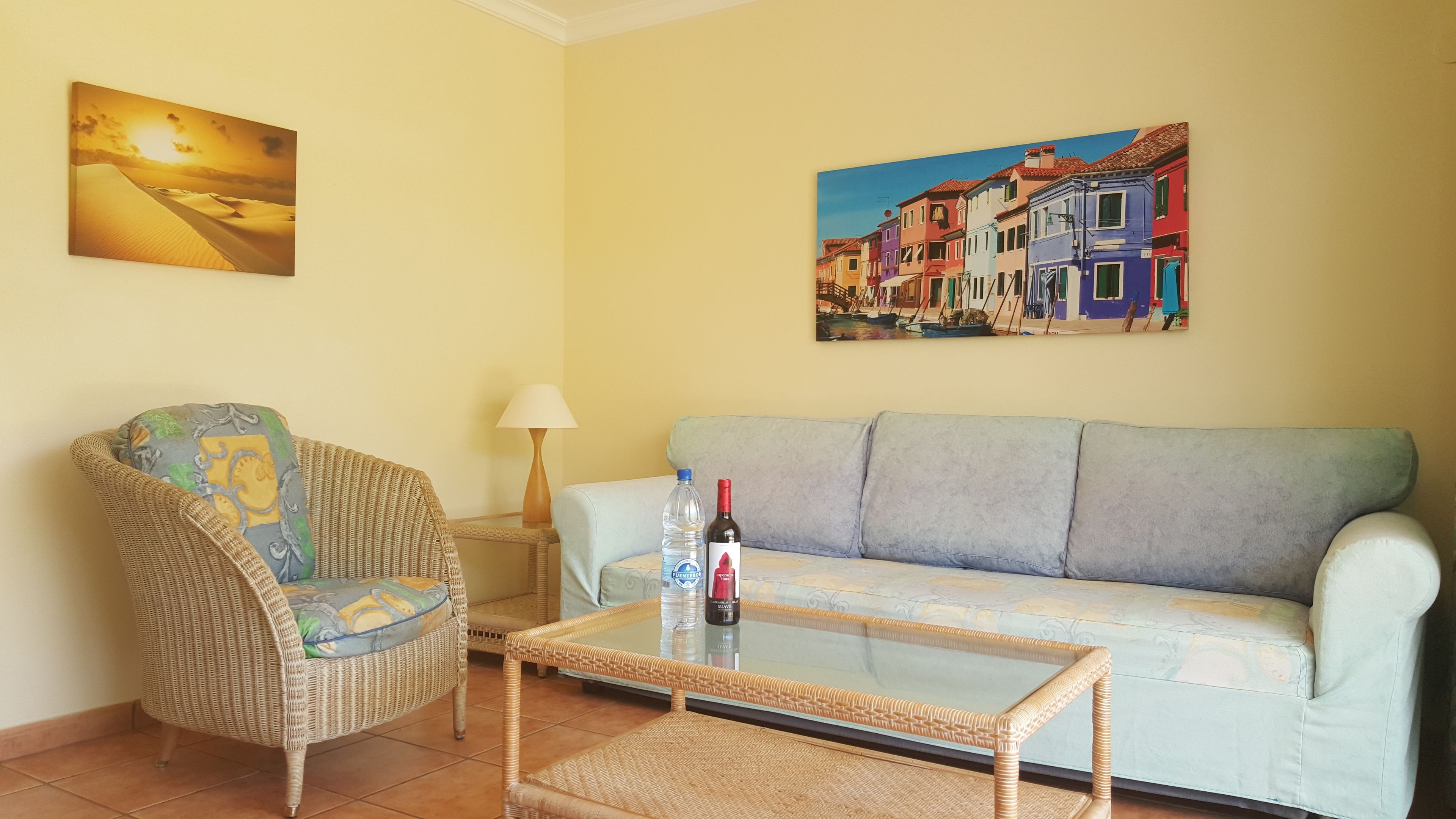 A living room with a wicker chair, a sofa, a glass coffee table, and two paintings on the wall.